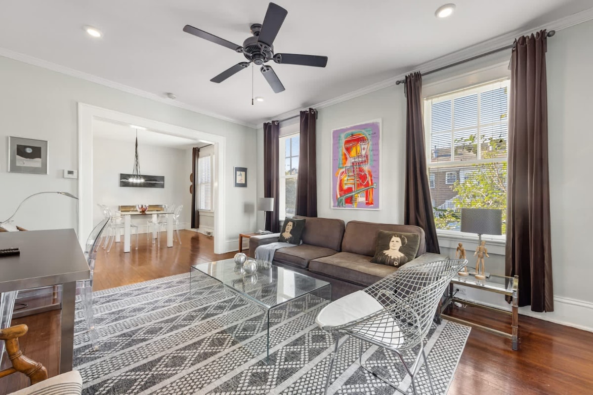 A contemporary living room showcases a dark upholstered sofa and a clear glass coffee table atop a patterned area rug. A ceiling fan is installed, while large windows with dark curtains allow natural light to fill the space. The adjacent dining area is visible in the background.