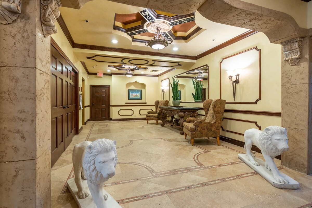 An inviting lobby area features elegant design with decorative details on the ceiling and walls. Stone floors are complemented by two lion sculptures flanking the entrance. Comfortable seating is available, alongside potted plants for a touch of greenery in the space.