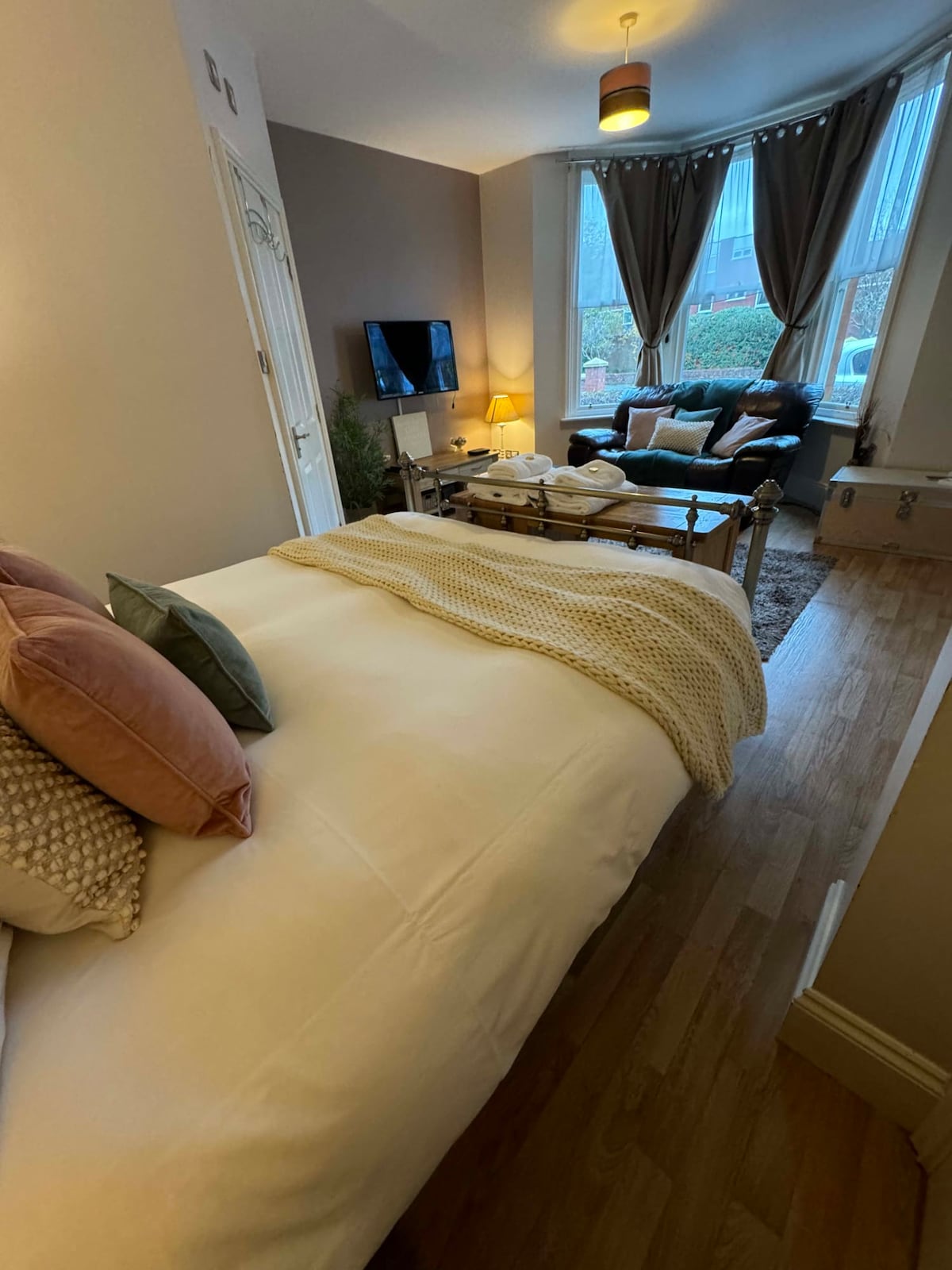 The studio features a spacious bed adorned with decorative pillows and a knitted throw. A cozy seating area with a leather sofa is visible by the window, which is framed by elegant curtains. A television is mounted on the wall, contributing to the inviting ambiance.