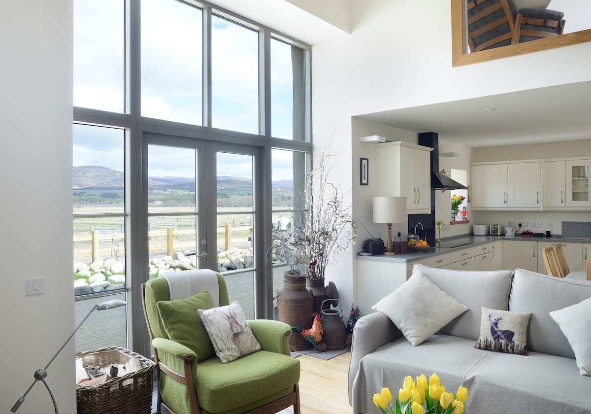 An open plan living space is presented, featuring large windows that allow natural light and picturesque views of the surrounding landscape. A comfortable green armchair and a light grey sofa are arranged around a central table. Decorative elements include a bouquet of yellow tulips and a basket, enhancing the inviting atmosphere.