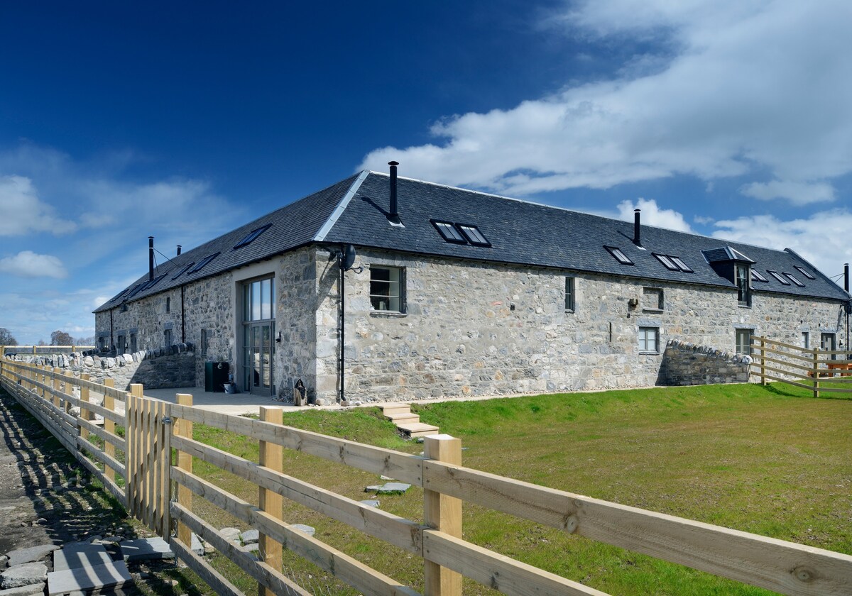 A stone-built lodge features a gabled roof and multiple windows, allowing natural light to brighten the interior. The exterior is complemented by a grassy area with a wooden fence, highlighting the rural setting and inviting views of the surrounding landscape.