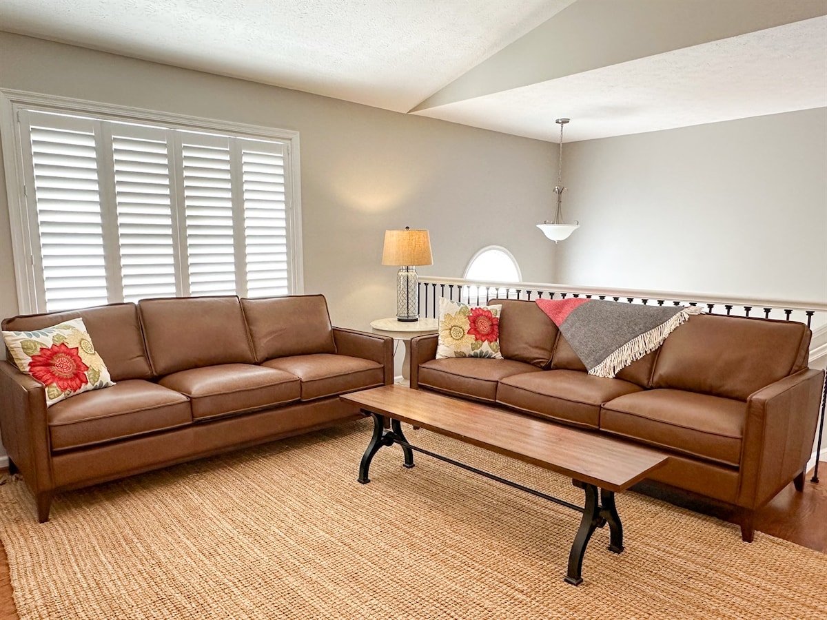 The living area features two brown upholstered sofas adorned with decorative pillows. A wooden coffee table rests in front, with a woven area rug covering the floor. Natural light enters through the shuttered windows, and a pendant light adds ambient illumination.