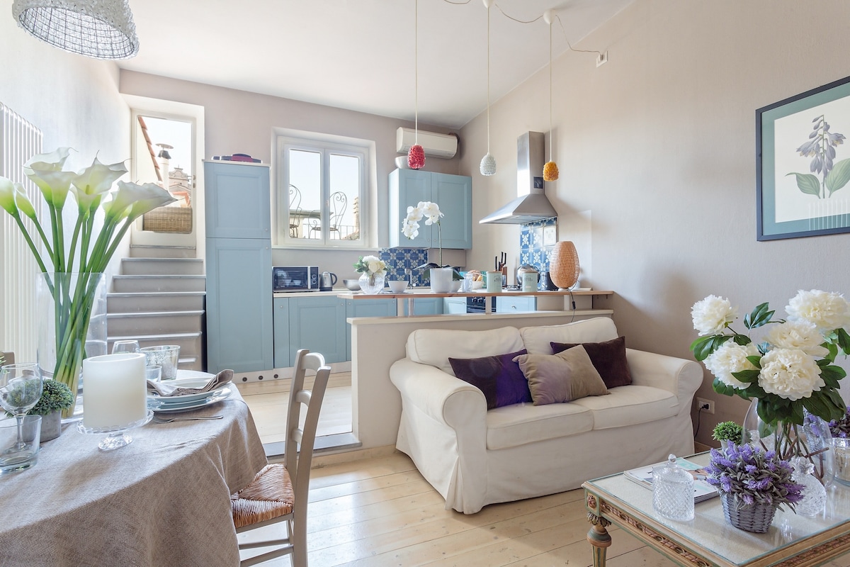 An inviting living area features a light-colored sofa adorned with cushions. A dining table, set with a neutral tablecloth, showcases fresh flowers. Blue cabinets are arranged in the kitchen area, while sunlight brightens the space through a large window, adding a warm ambiance.
