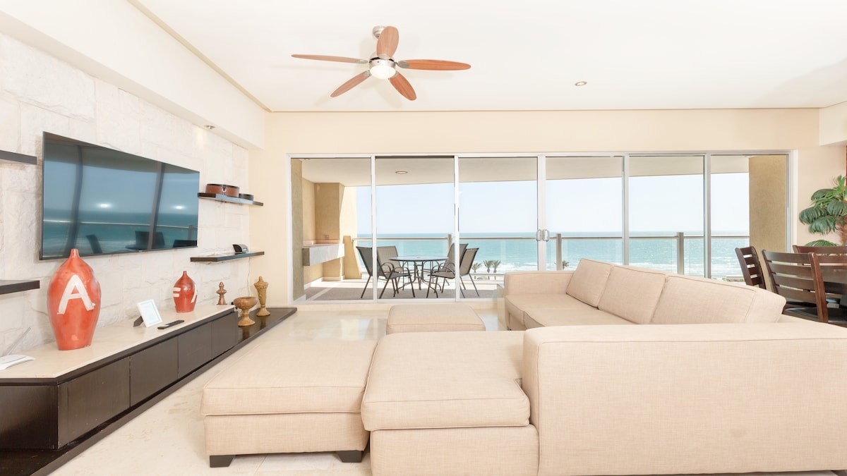 A spacious living area features a light-colored sectional sofa facing a mounted flat-screen TV. Large glass doors provide a view of the ocean and access to a private terrace with outdoor seating. A ceiling fan is installed for comfort, enhancing the bright, airy feel of the space.