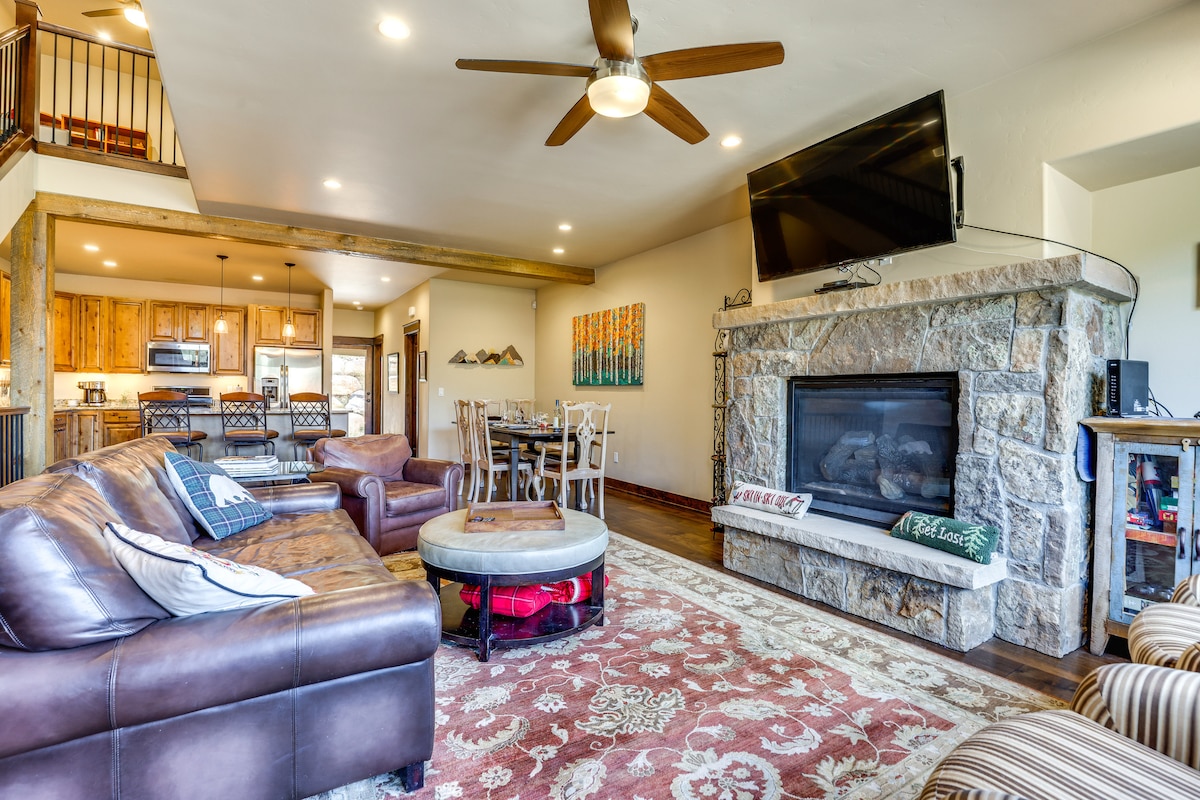The spacious living area features a combination of leather seating arrangements centered around a stone fireplace. A large television is mounted above the fireplace, and natural light floods the space. A dining area is visible in the background, surrounded by warm wooden accents.
