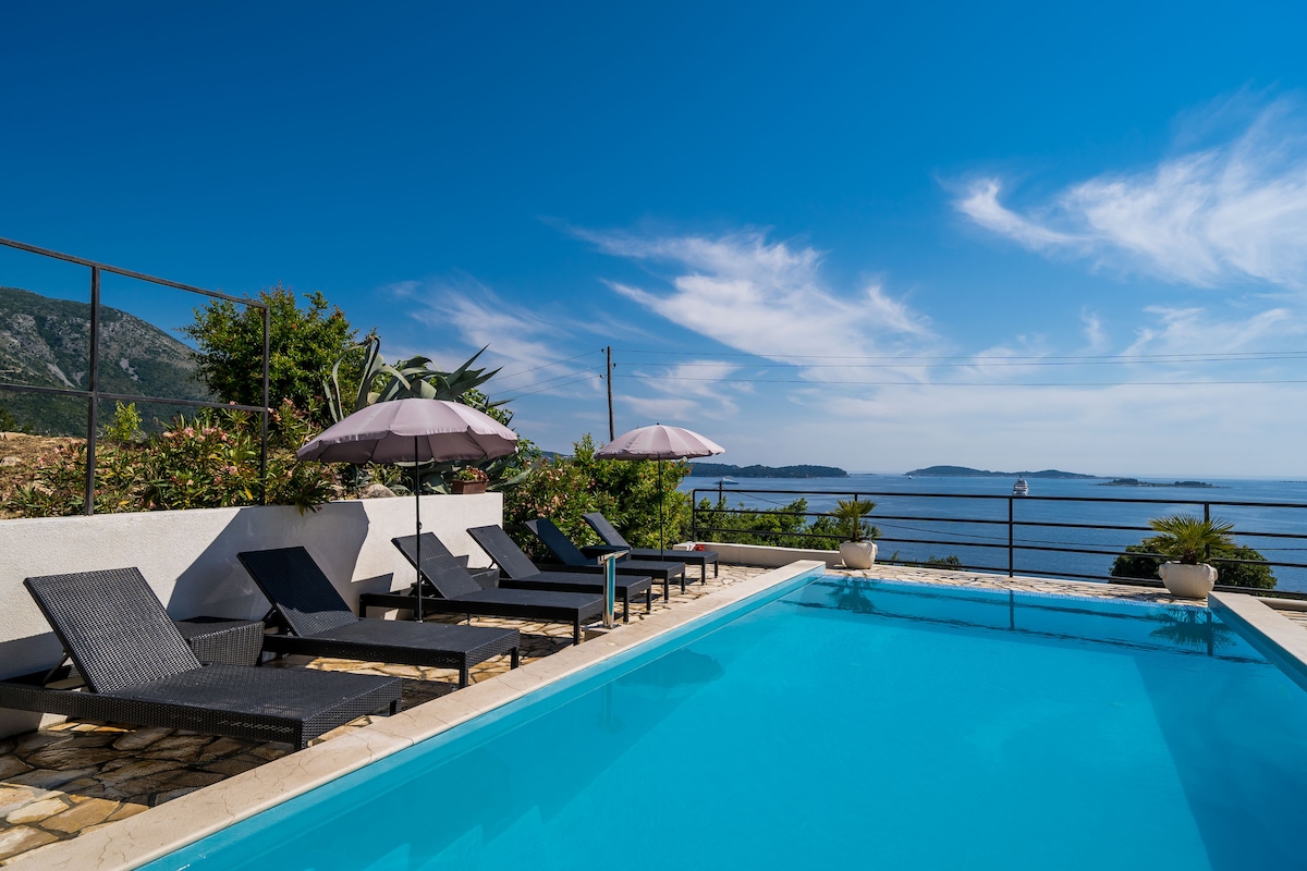 An outdoor seasonal swimming pool is surrounded by a spacious sun terrace. Several lounge chairs are arranged with matching umbrellas, offering shade. Views of the sea and distant islands are visible beyond the pool, under a clear blue sky with scattered clouds.