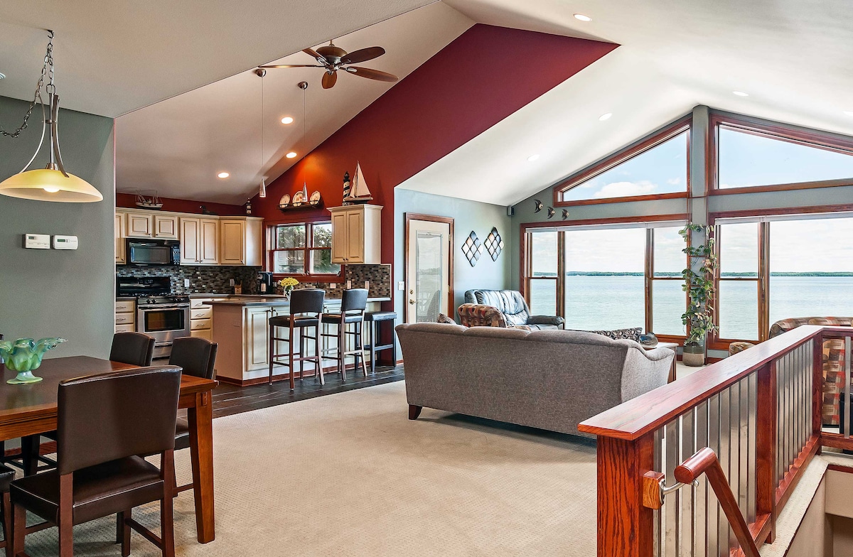 The open-plan living space features large windows overlooking the lake, inviting natural light to flow in. A dining area with a wooden table sits beside a modern kitchen, equipped with light cabinetry. A comfortable seating area is arranged with a sofa and an inviting view of the water.