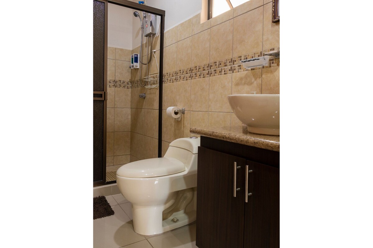 A modern bathroom is presented, featuring a glass-enclosed shower with a handheld showerhead. A sleek vanity with a round sink complements the neutral-toned tiles. A towel bar and toilet paper holder are conveniently placed, ensuring accessibility.