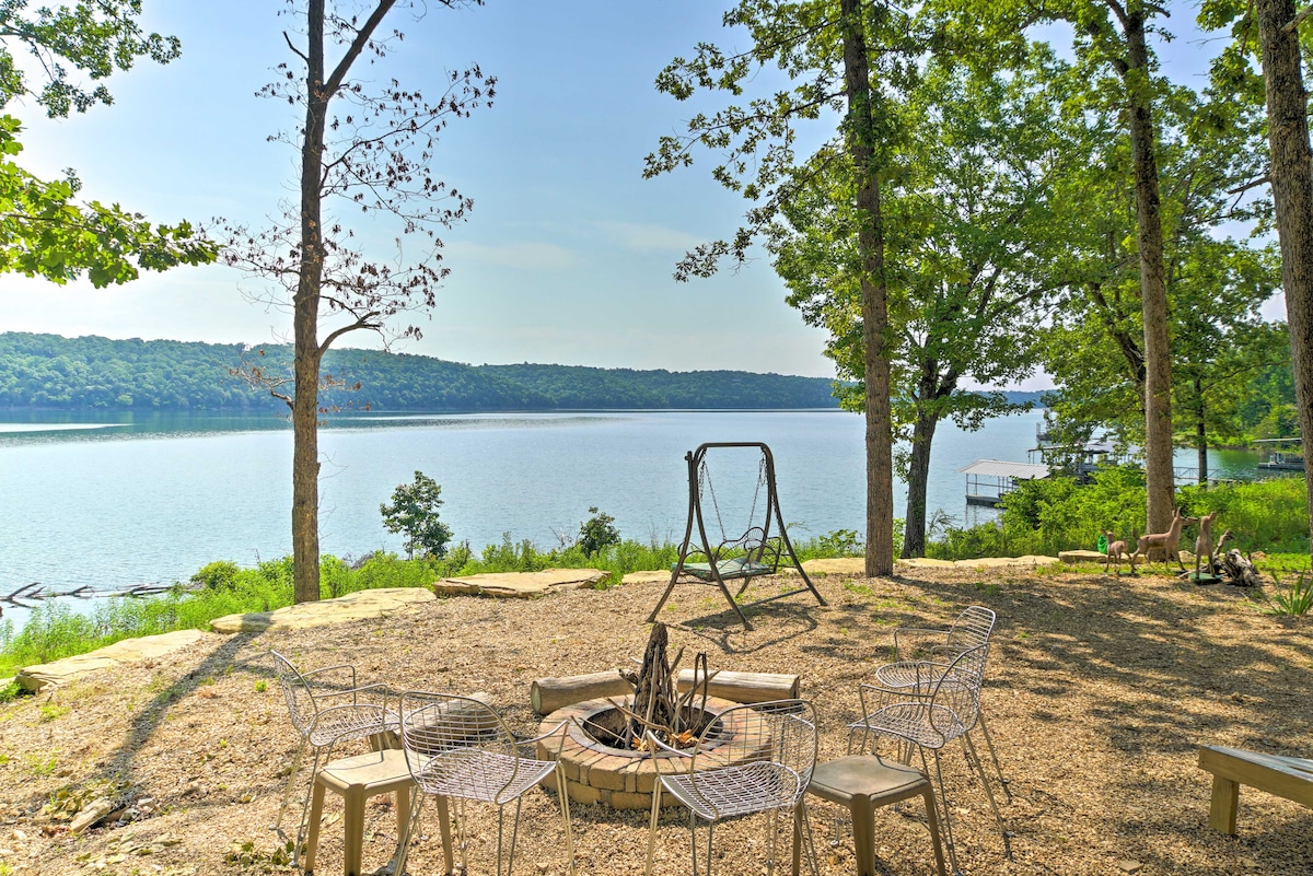 A stone fire pit is surrounded by wireframe chairs, offering a scenic view of Bull Shoals Lake. A wooden swing hangs nearby under the shade of trees, overlooking calm waters and the lush green shoreline.