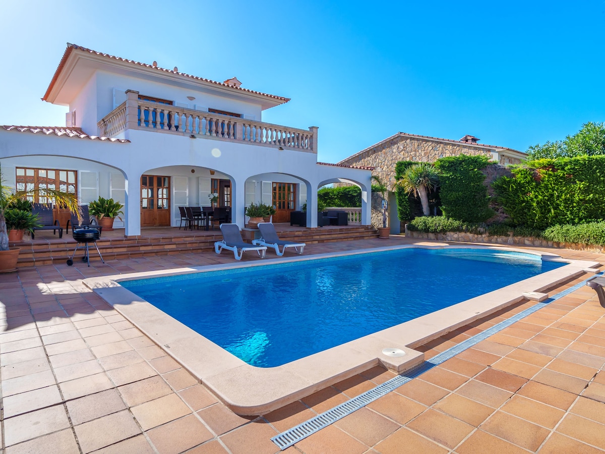 A two-story house is depicted with a spacious terrace overlooking a swimming pool. The pool area features lounge chairs and a barbecue, surrounded by greenery. Large glass doors allow natural light into the house, contributing to a bright and open atmosphere.