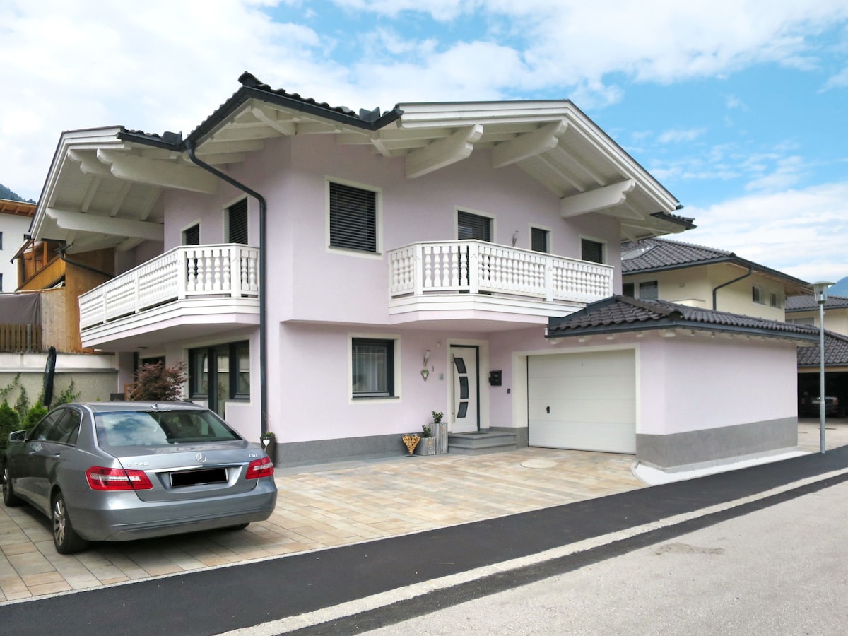 The exterior of a two-family house is presented, featuring a light pink facade and decorative balcony railings. A gray car is parked in front of the tiled entrance. The building includes a garage and showcases a blend of modern design with traditional alpine elements under a cloudy blue sky.