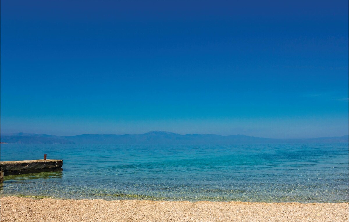 A serene beach is viewed with calm, clear waters gently lapping against the sandy shore. The horizon features a seamless transition of blue from the water to the sky, with distant mountains faintly visible. A wooden pier extends into the water, offering a peaceful spot for relaxation.
