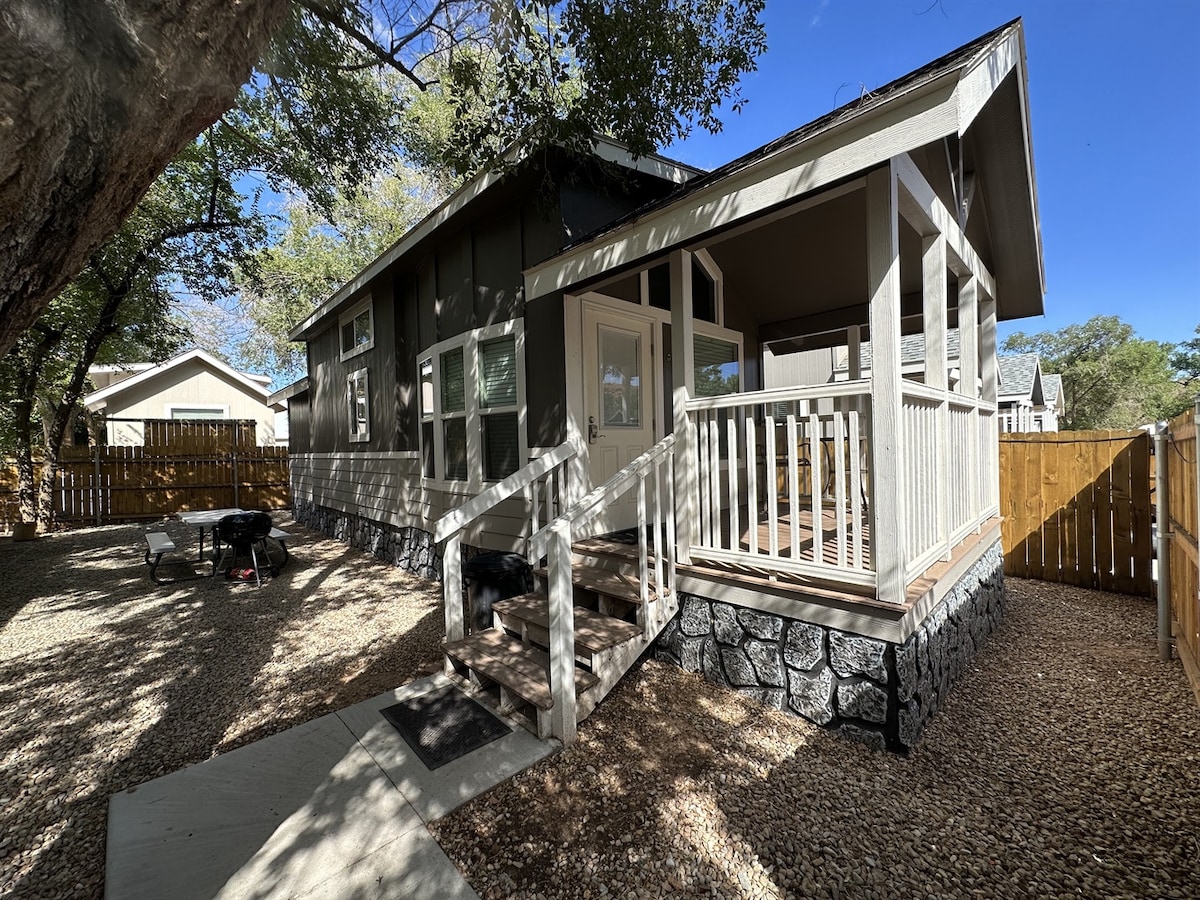 A charming tiny home is surrounded by a well-maintained yard, featuring a gravel driveway and a wooden patio with railing. Sunlight filters through the trees, casting playful shadows. The exterior showcases dark siding and light trim, complemented by a fenced outdoor area for privacy.