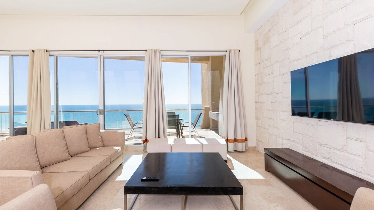 A spacious living room is highlighted by large glass doors that offer panoramic views of the ocean. A neutral-colored sectional sofa surrounds a dark coffee table, while natural light fills the area. A flat-screen TV is mounted on the wall, enhancing the modern design.