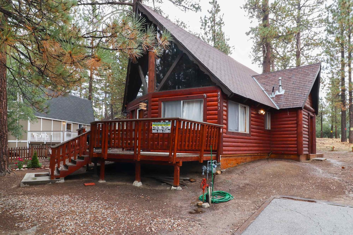 A rustic cabin constructed from logs features a sloped roof and large windows allowing natural light to fill the interior. A wrap-around deck provides accessibility and outdoor space, while the surrounding trees create a natural, serene environment.