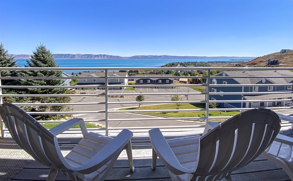 A private balcony features two white adirondack chairs, providing a perfect spot for relaxation. The expansive lake view is highlighted, showcasing clear blue water and distant hills under a bright sky.
