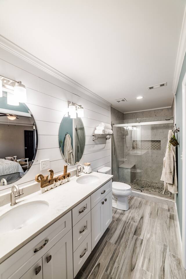 The bathroom showcases a dual vanity with circular mirrors and modern lighting. A walk-in shower, enclosed in glass, features elegant marble tiling. The space is completed with light-colored wood-like flooring and a calming color scheme, creating a fresh and clean atmosphere.