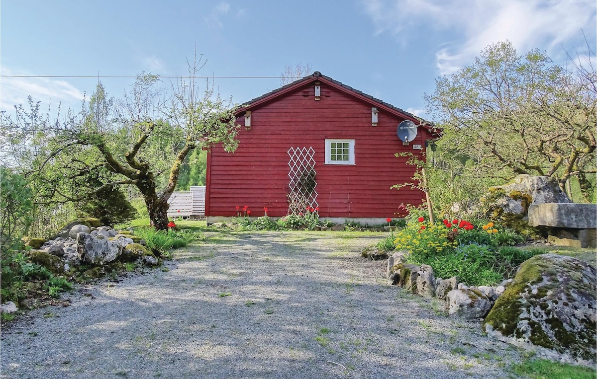 A red cottage is set amidst a well-maintained garden, with colorful flowers and lush greenery surrounding the stone pathway. The building features a small window and decorative trellis, providing a welcoming focal point in the serene natural environment.