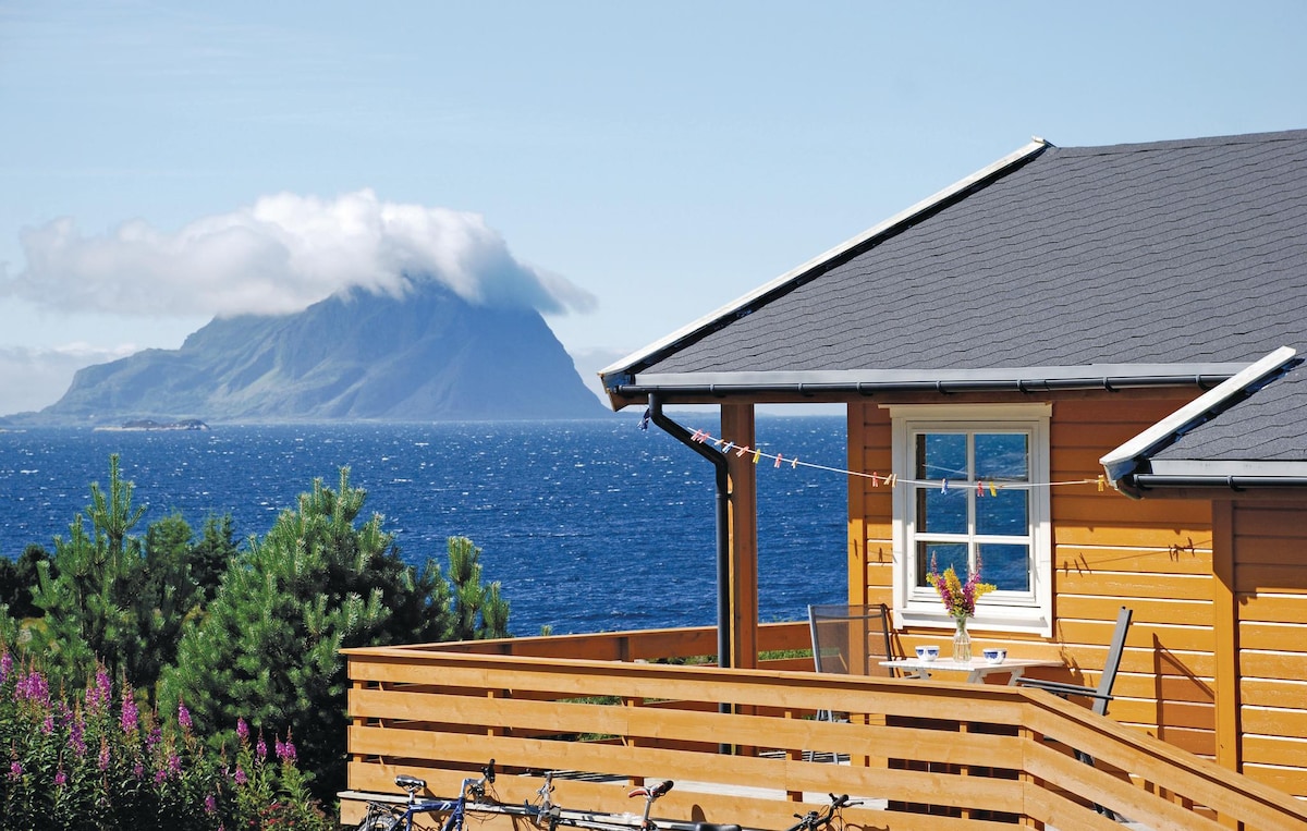An orange wooden house is depicted with a large terrace that features a table and chairs. A mountain with clouds forms the background over the sea, while greenery and purple flowers are visible in the foreground.
