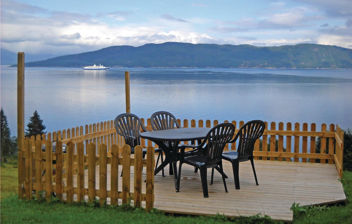 A wooden deck featuring a table surrounded by four black chairs overlooks calm waters of the fjord. In the background, hills rise gently against a cloudy sky, with a boat visible in the distance, enhancing the serene outdoor setting.