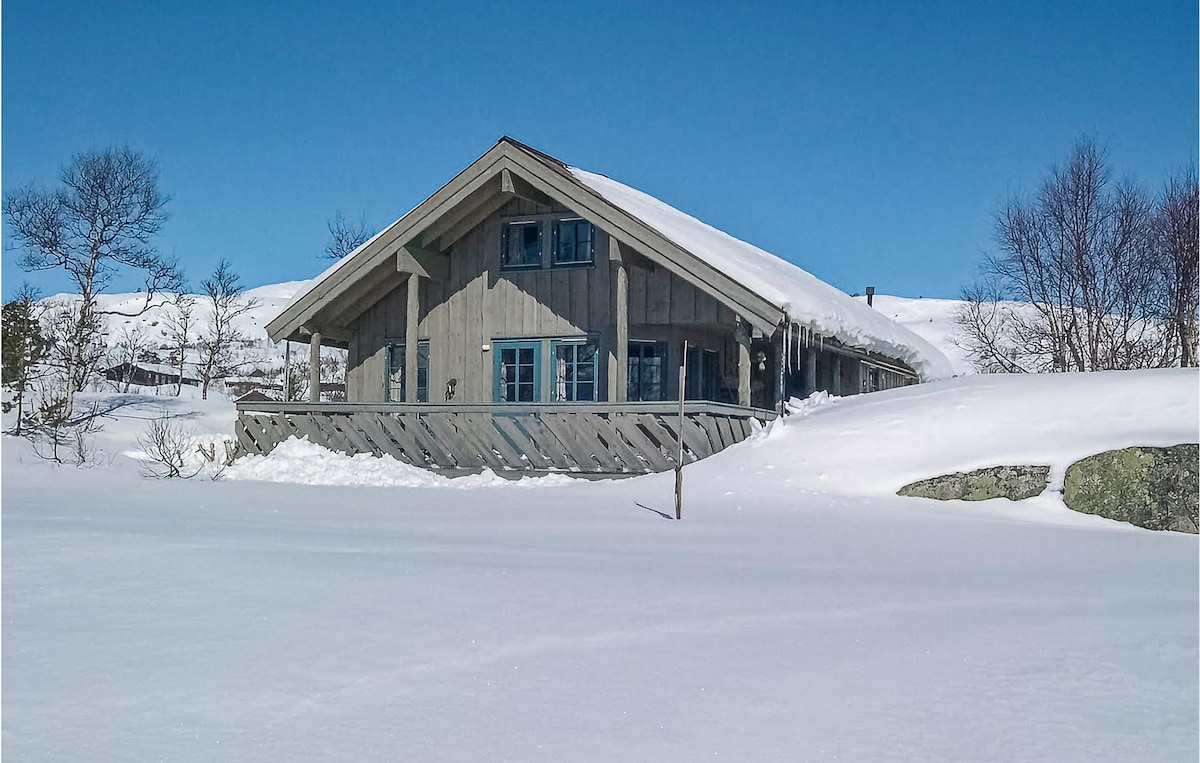 The holiday home features a wooden exterior with a sloped roof, set against a clear blue sky. Snow covers the surrounding landscape, creating a serene atmosphere. Large windows are visible, reflecting the natural light, while the decking offers potential for outdoor relaxation.