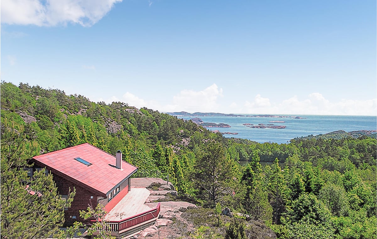 A charming vacation home is nestled among lush greenery, featuring a red roof and panoramic views of the sea. The vibrant landscape includes a mix of trees and rocky terrain, with distant islands visible on the horizon under a clear blue sky.