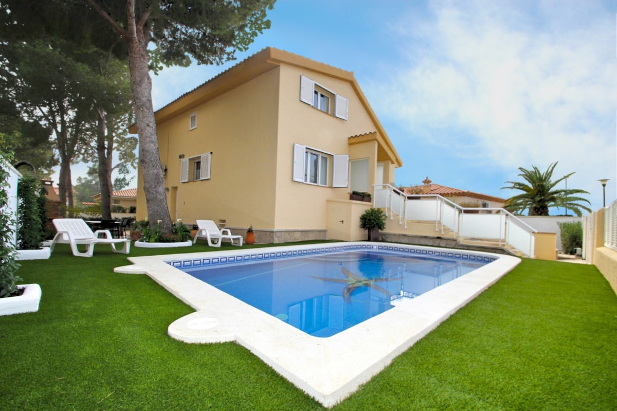 A private pool is surrounded by well-maintained grass and features a shallow entry area. The villa is situated nearby, showcasing a two-story design with white window shutters. Lush greenery and palm trees enhance the outdoor space, creating a tranquil setting.