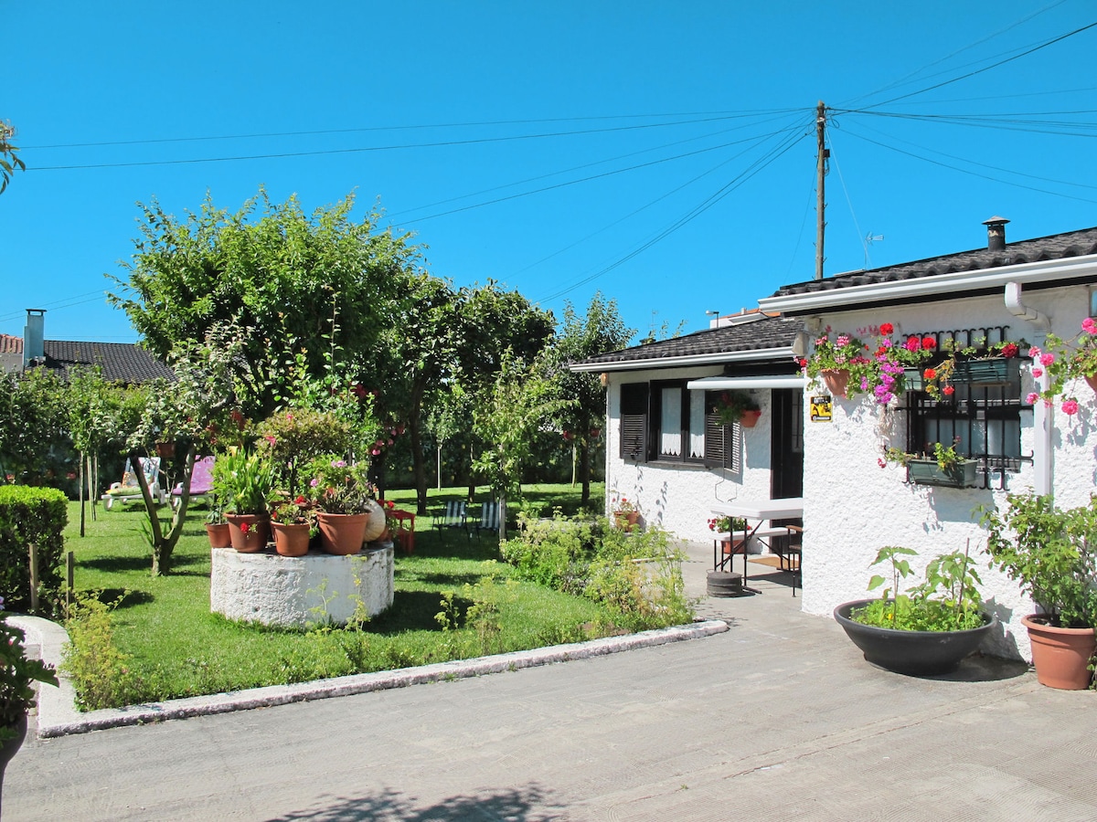 A well-maintained bungalow is nestled in a garden setting, featuring a neatly trimmed lawn and various plants. Nearby, a round flower bed with potted plants adds color and interest. The exterior of the bungalow is complemented by large windows and outdoor seating areas for relaxation.