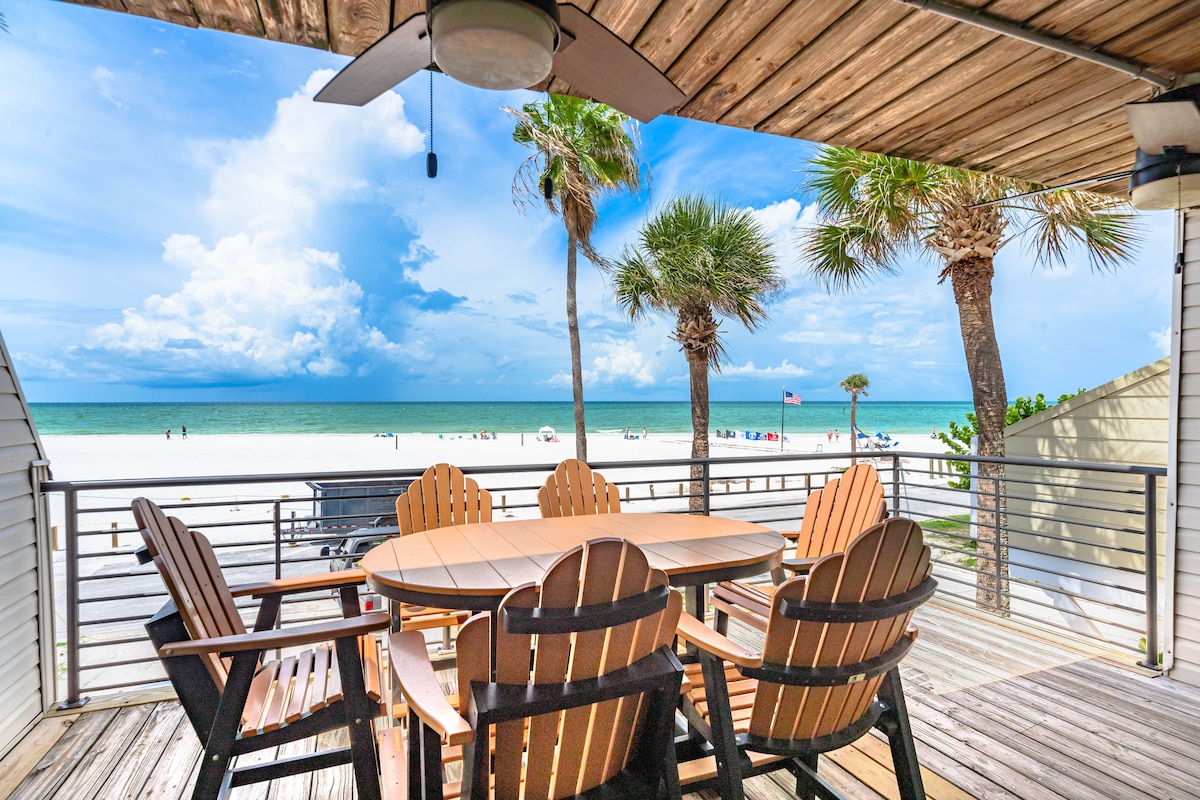 A spacious outdoor deck is depicted, featuring a circular wooden dining table surrounded by sturdy chairs. Lush palm trees frame the view, which overlooks the sandy beach and turquoise waters. The sky is bright with scattered clouds, enhancing the seaside atmosphere.