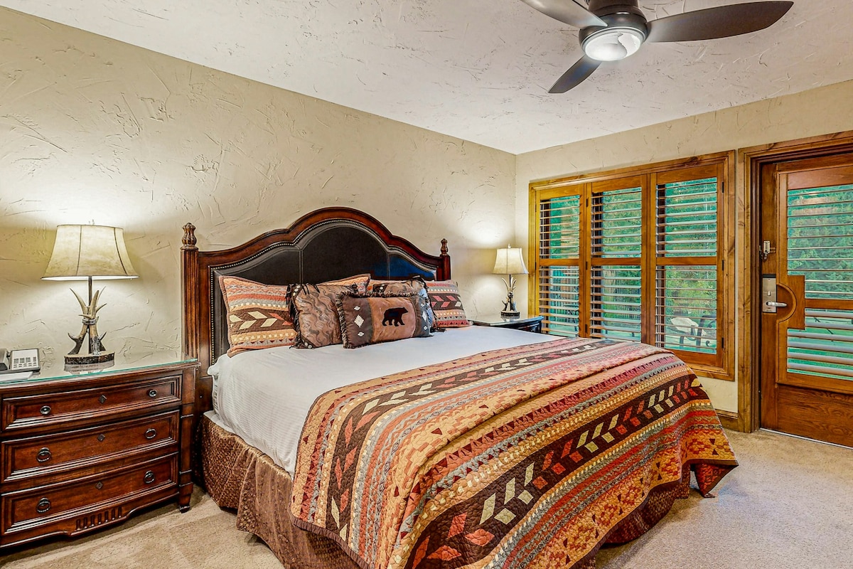 A spacious bedroom features a king bed adorned with a vibrant patterned blanket. Two bedside lamps provide soft lighting alongside a wooden nightstand. The room includes window shutters that allow for natural light, with a door leading outside, complementing the cozy ambiance.