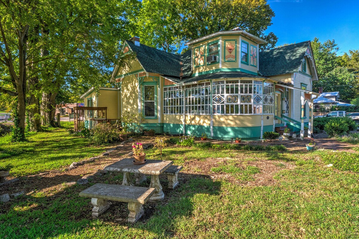 A charming Victorian house is framed by mature trees and manicured landscaping. A cozy stone table and benches are positioned in the grassy yard, offering a serene spot for relaxation. The home features a unique architectural design with colorful accents and large windows that invite natural light.