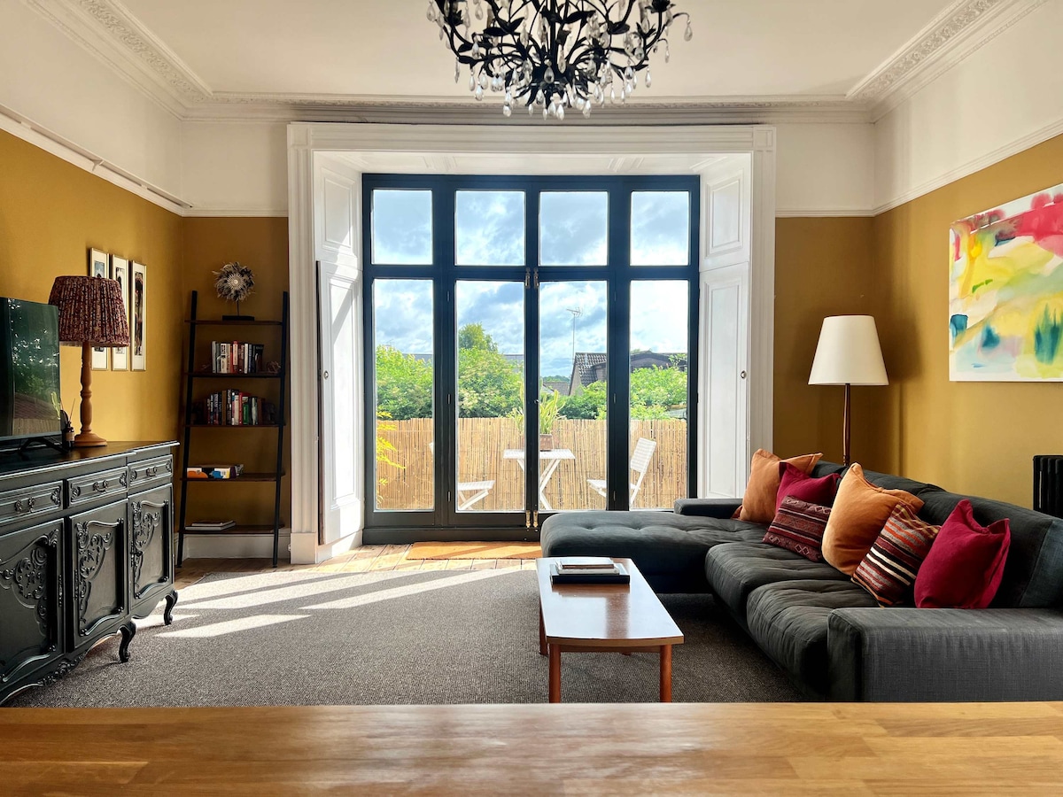A spacious living area is illuminated by natural light flooding through large, double-aspect sash windows. A charcoal sectional sofa adorned with colorful pillows faces a low coffee table. A decorative cabinet and television are positioned against a warm-colored wall, while a balcony with outdoor seating is visible beyond.