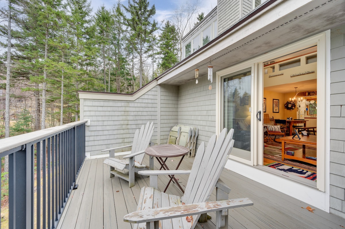 An outdoor deck area features a circular wooden table surrounded by four Adirondack chairs. The deck is bordered by trees, providing a natural view. Sliding glass doors lead into a cozy interior space, creating a seamless transition between indoor and outdoor living.