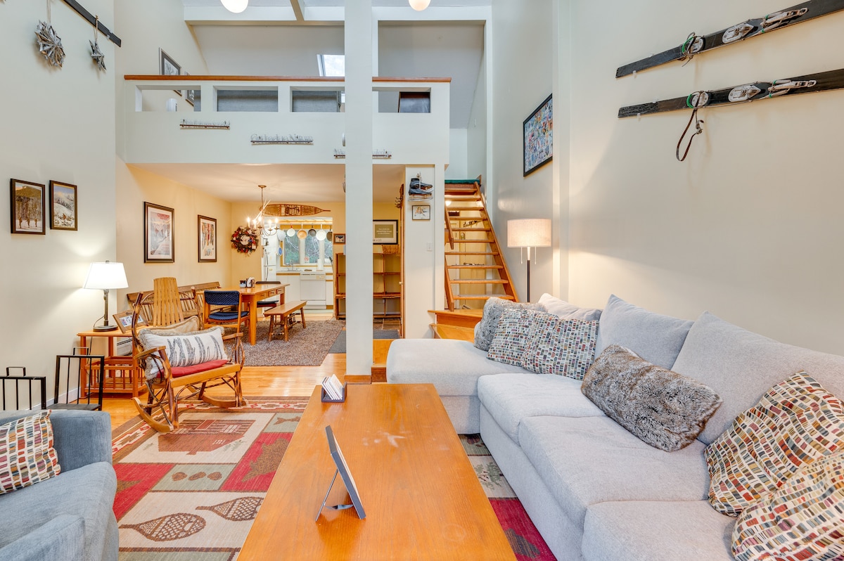 The open living space features a light and airy design, complemented by a large sectional sofa and a wooden coffee table. Ski decor adorns the walls, alongside framed art. A staircase leads to the upper level, while a cozy dining area can be seen in the background.