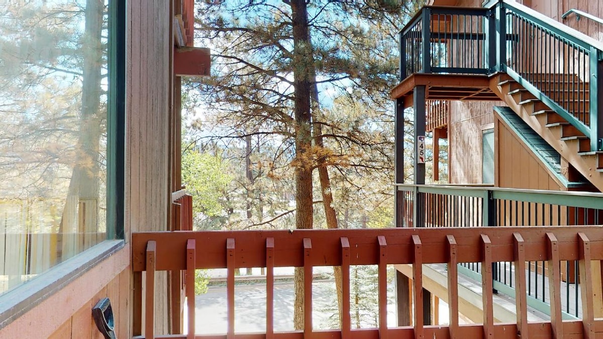 A view from the balcony reveals a wooden railing overlooking a tree-lined landscape. Staircases with green railings provide access to upper levels, while large windows facilitate natural light and connect the indoor and outdoor environments.