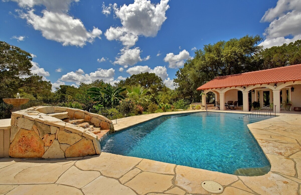 A serene pool area is framed by natural stone landscaping and vibrant greenery. The inviting water reflects the clear blue sky dotted with fluffy clouds. Surrounding the pool, a spacious patio provides ample lounging space, while a charming covered area is visible in the background.