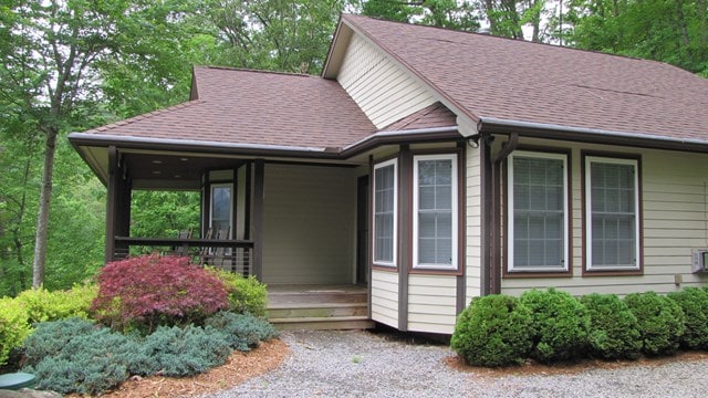 The exterior of the cottage is framed by lush greenery, featuring a welcoming front porch with multiple entry points. Large windows allow natural light to fill the interior, and neatly landscaped bushes and shrubs enhance the entrance.