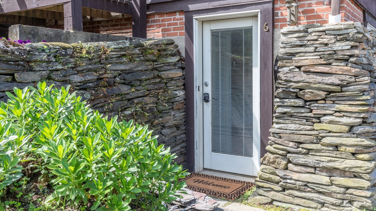 The entrance to the studio is framed by rustic stone walls and surrounded by lush greenery. A glass door with a keypad lock leads to the interior, while a welcome mat is positioned at the threshold.