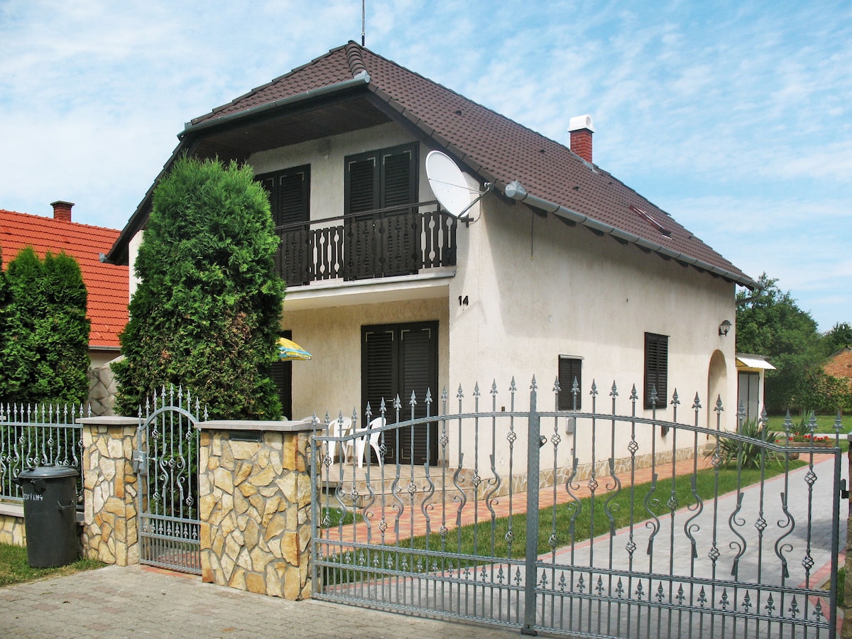 A two-story house is presented with a light-colored facade and dark shutters. The gated entrance leads to a well-maintained garden. A balcony is visible on the upper level, and a few chairs are positioned in the outdoor area. Rolling green lawn surrounds the property.