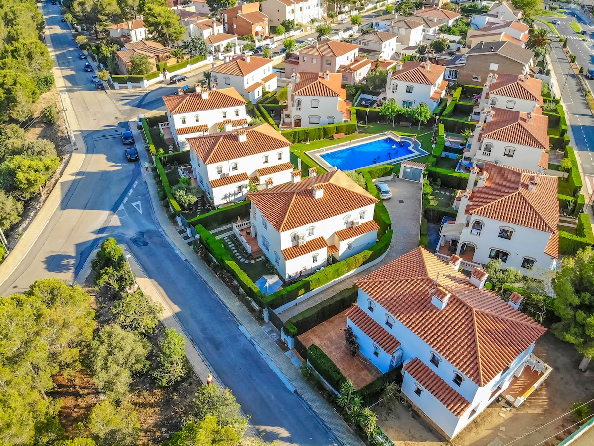 An aerial view captures a group of two-story townhouses surrounded by well-maintained gardens and pathways. A communal swimming pool is prominently featured in the center, inviting for relaxation. Paved streets and neatly arranged homes highlight the organized layout of the neighborhood.