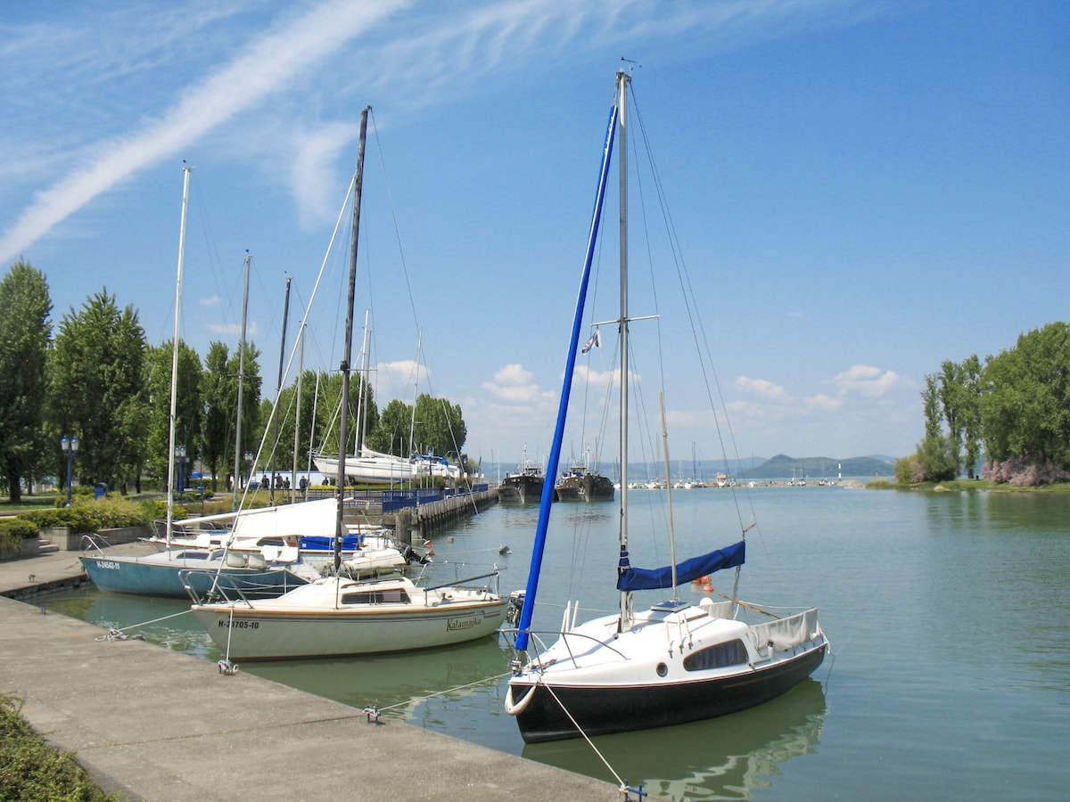 Two sailboats are moored at a quiet marina, surrounded by lush trees along the water's edge. A clear blue sky with wispy clouds reflects on the calm lake surface. The dock is visible, offering a serene atmosphere by the water.