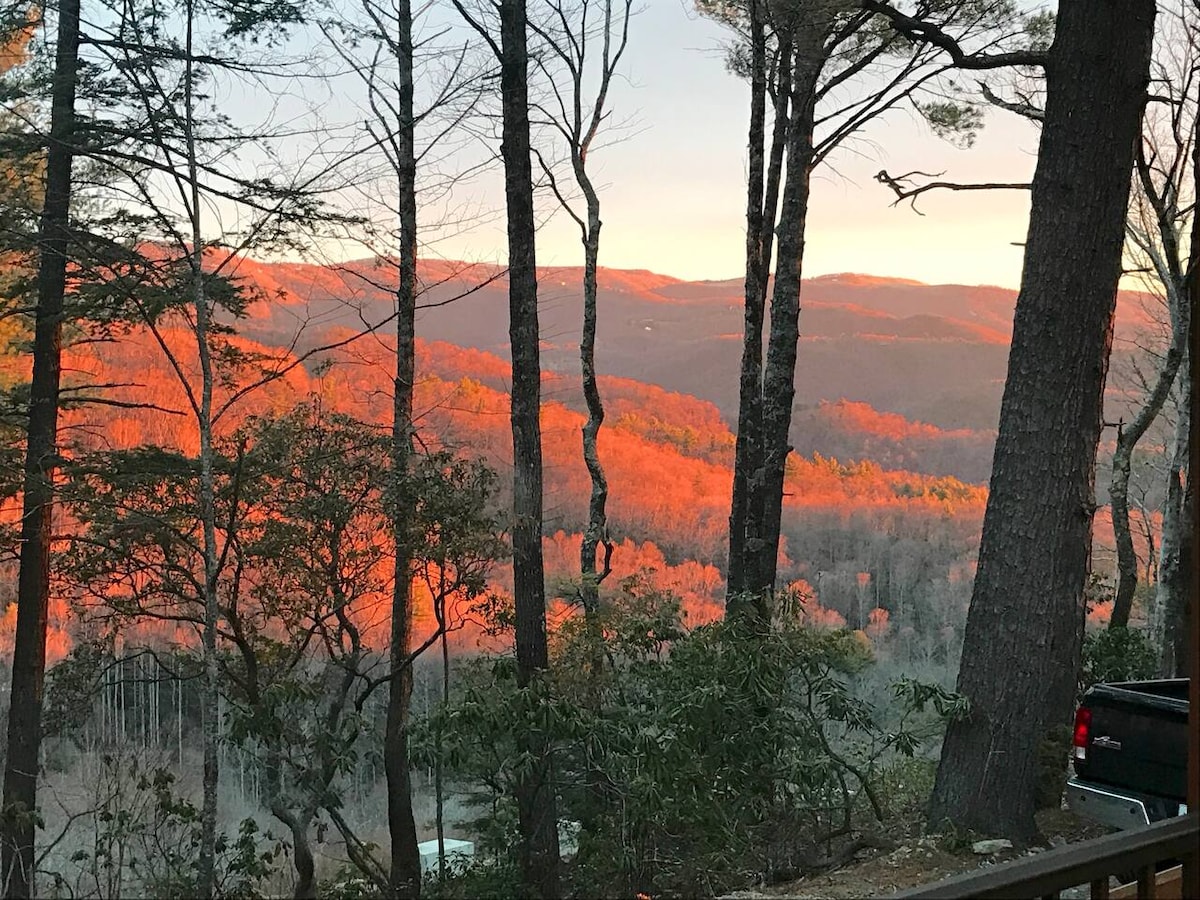 The image captures a vibrant sunset illuminating the mountainside, with trees in the foreground framing the view. The warm hues of orange reflect off the autumn foliage, showcasing the natural beauty of the surrounding landscape.