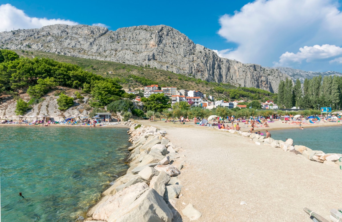 A tranquil beach scene is captured, highlighting a sandy shoreline bordered by clear turquoise water. A rocky path leads into the sea, with sunbathers and families enjoying their time on the beach. Lush greenery and rocky hills rise in the background, creating a scenic landscape.