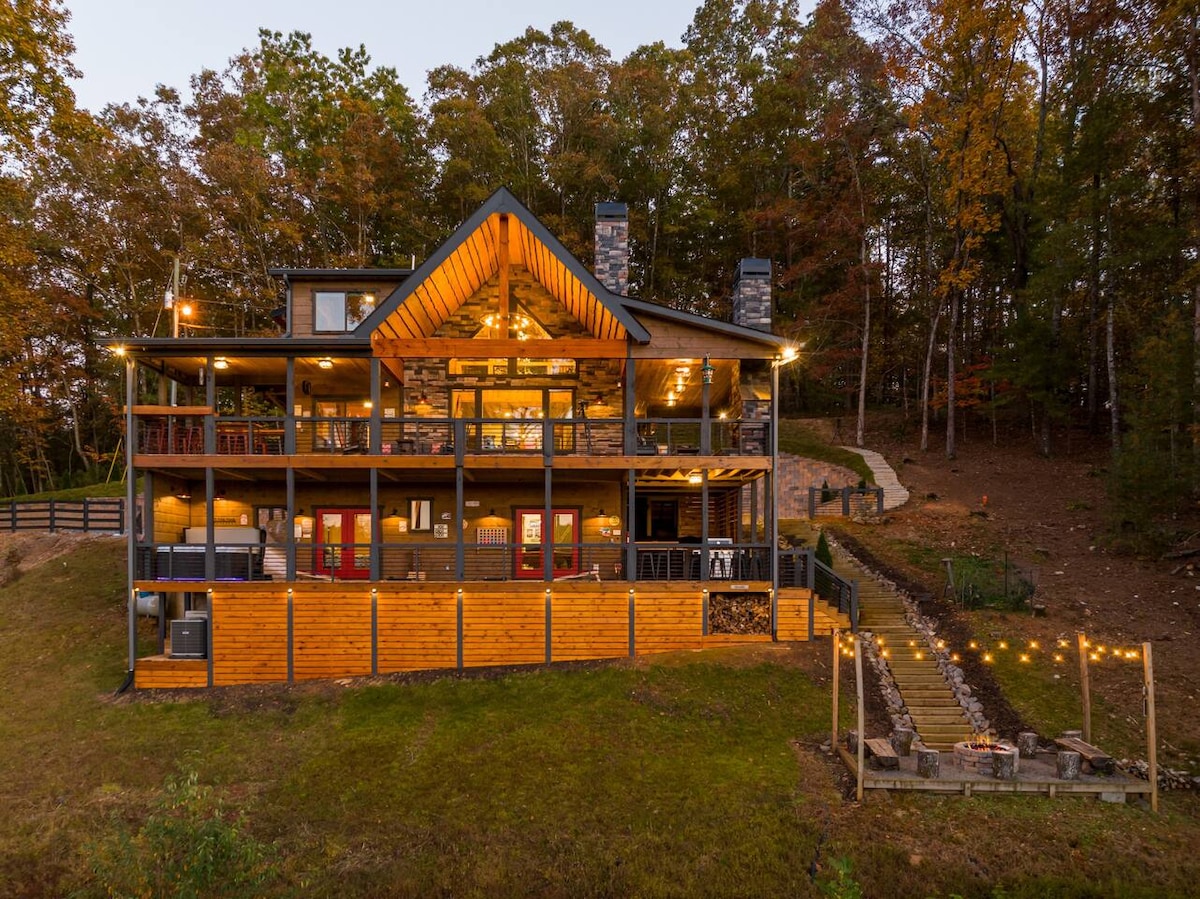 The cabin's exterior is highlighted by natural wood and stone accents, with multiple levels and expansive decks. Warm lights illuminate the structure, while trees with autumn foliage surround the property, creating a welcoming and serene atmosphere.