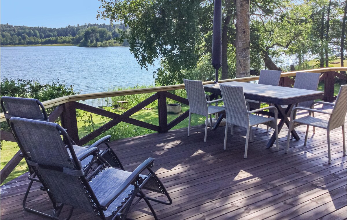 A large wooden terrace overlooks the lake, featuring a dining table surrounded by six chairs. Two reclining loungers are positioned for relaxation. Lush greenery borders the space, and the calm water reflects the surrounding nature.