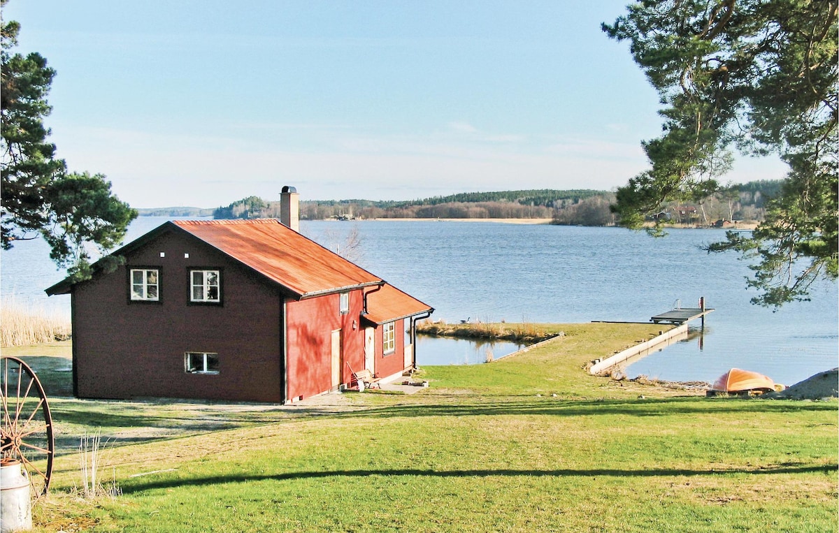 A red cottage with a sloped roof is set against a serene lake backdrop. The property features lush green grass and trees, with a wooden pier extending into the calm water. A small boat rests on the shore, enhancing the peaceful scene.