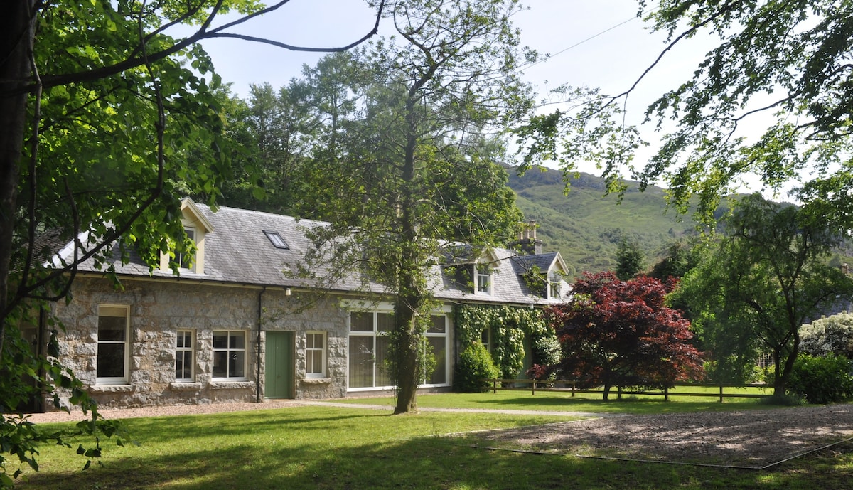A stone cottage with a slate roof is framed by lush greenery. Large windows allow natural light to fill the interior, while a vibrant tree provides a splash of color in the garden. The surrounding landscape features gentle hills in the background.