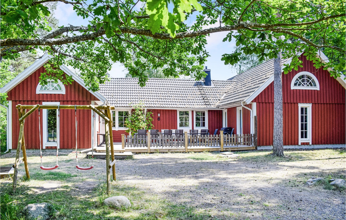 The red wooden house is set amidst greenery, featuring a welcoming porch with seating arrangements. A swing set is positioned in the foreground on a gravel surface, enhancing the outdoor space. The structure showcases large windows, allowing natural light to enter.