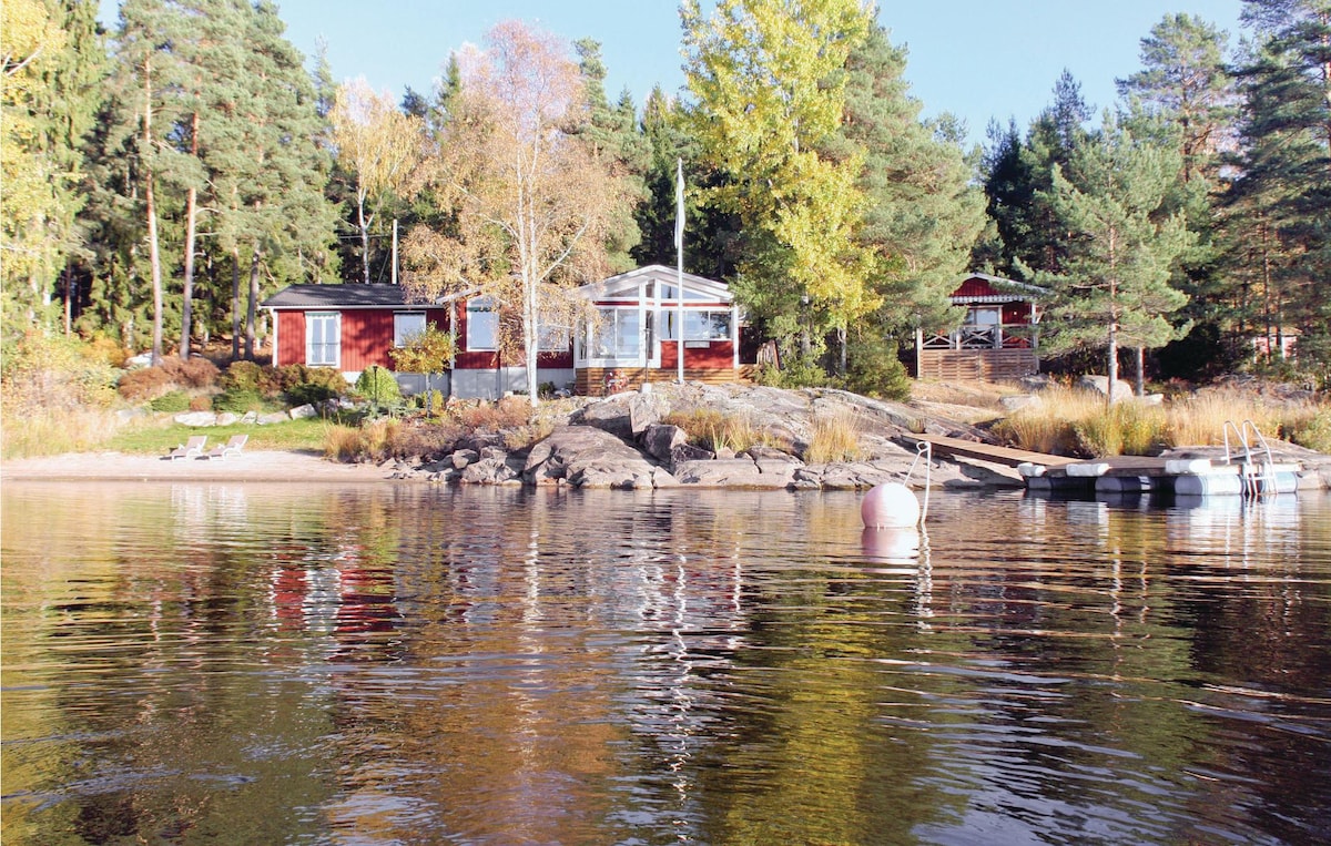 A vacation home is positioned along the shoreline, featuring wooden exteriors in red and dark tones. The reflection of the trees and cottage can be seen in the calm water. A small dock extends into the water, accompanied by a white buoy, creating a serene lakeside view.