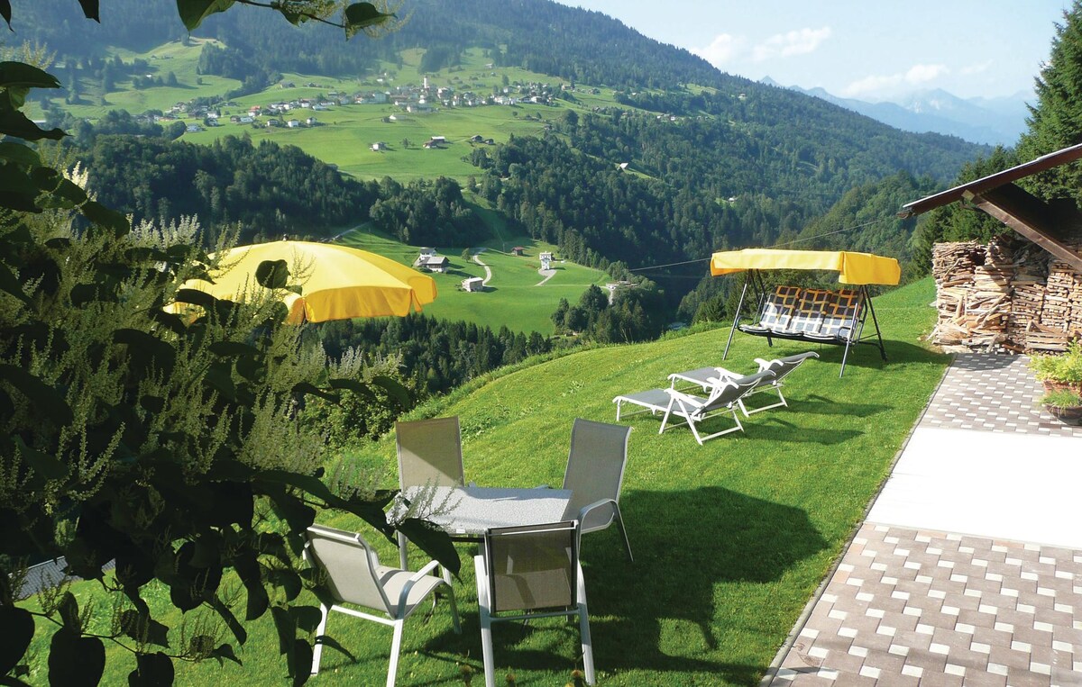 An open garden space is featured, showcasing sun loungers and patio furniture set against a backdrop of lush green hills. Two large yellow umbrellas provide shade, while a view of rolling valleys and distant mountains extends beyond the garden area.